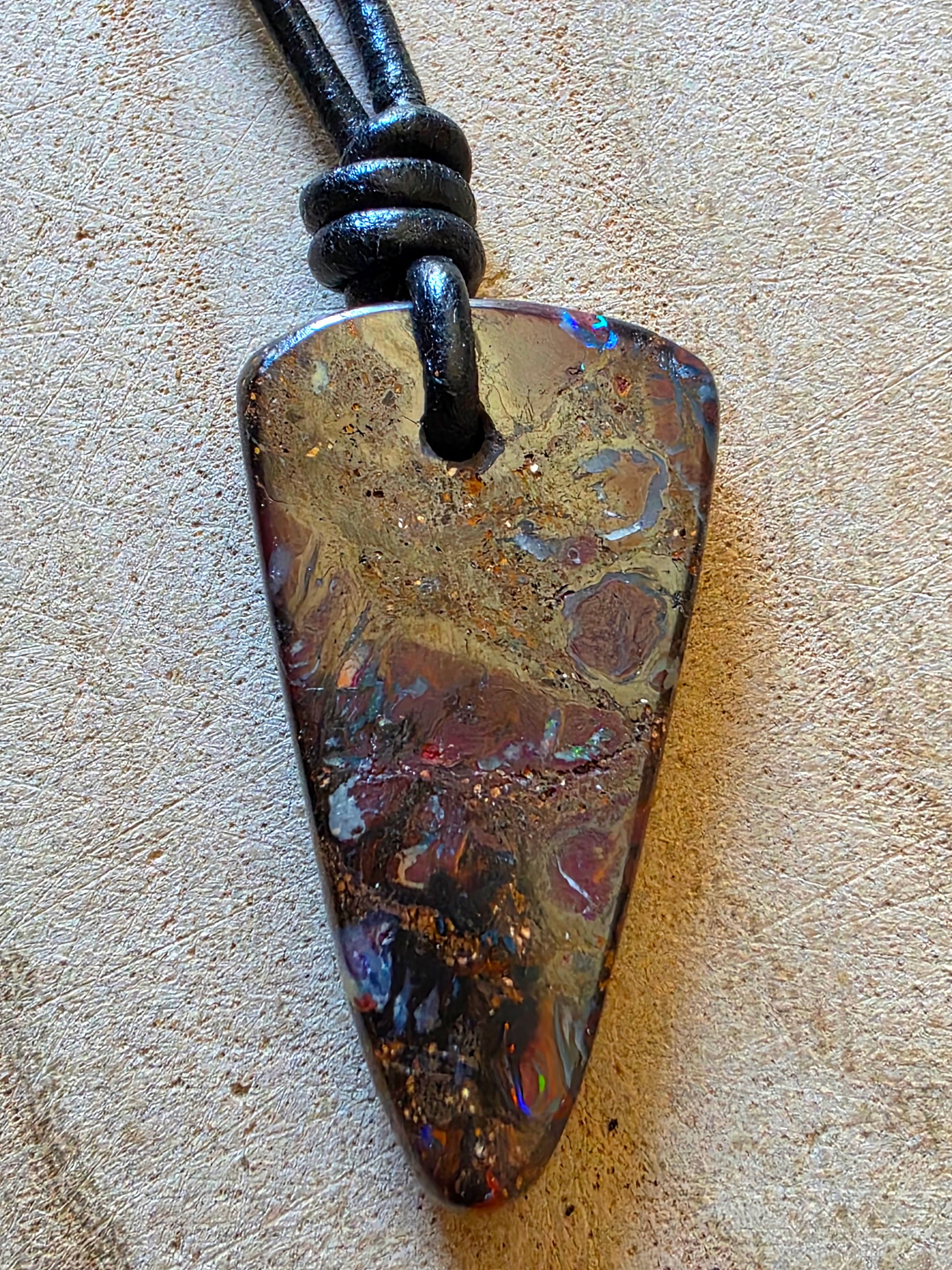 A natural opal pendant necklace with a triangle-shaped stone on a black cord, displayed on a textured beige surface.