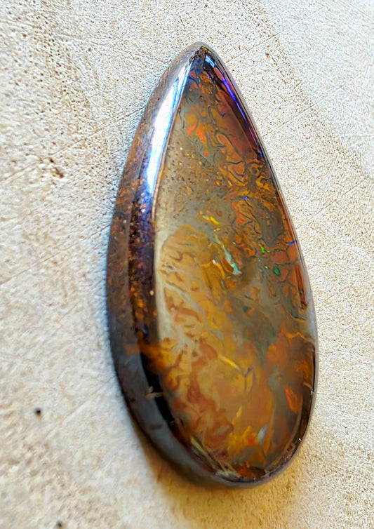 A teardrop-shaped cabochon opal with swirling patterns of amber, gold, brown, and iridescent blue, resting on a textured beige surface.