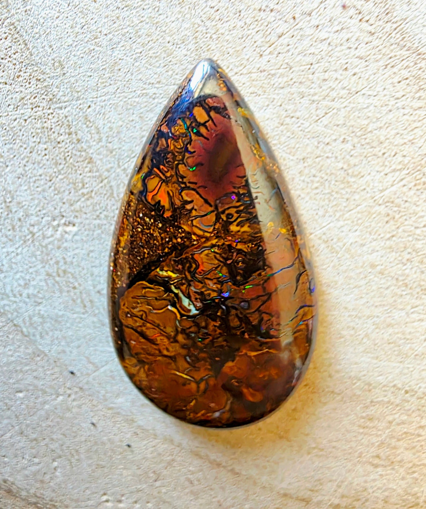 A teardrop-shaped opal cabochon featuring intricate golden, amber, and dark brown iridescent inclusions, resting on a textured beige surface.