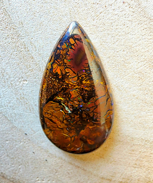 A teardrop-shaped opal cabochon featuring intricate golden, amber, and dark brown iridescent inclusions, resting on a textured beige surface.