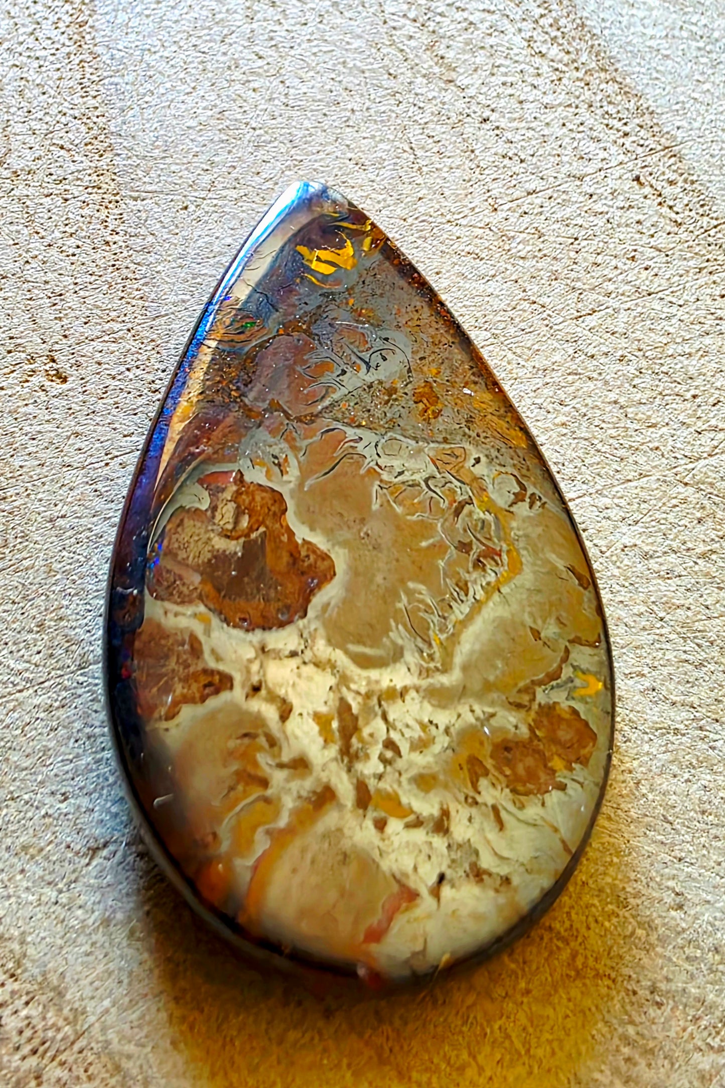 A teardrop-shaped cabochon opal featuring a marbled pattern of brown, beige, and gold, resting on a textured beige surface.