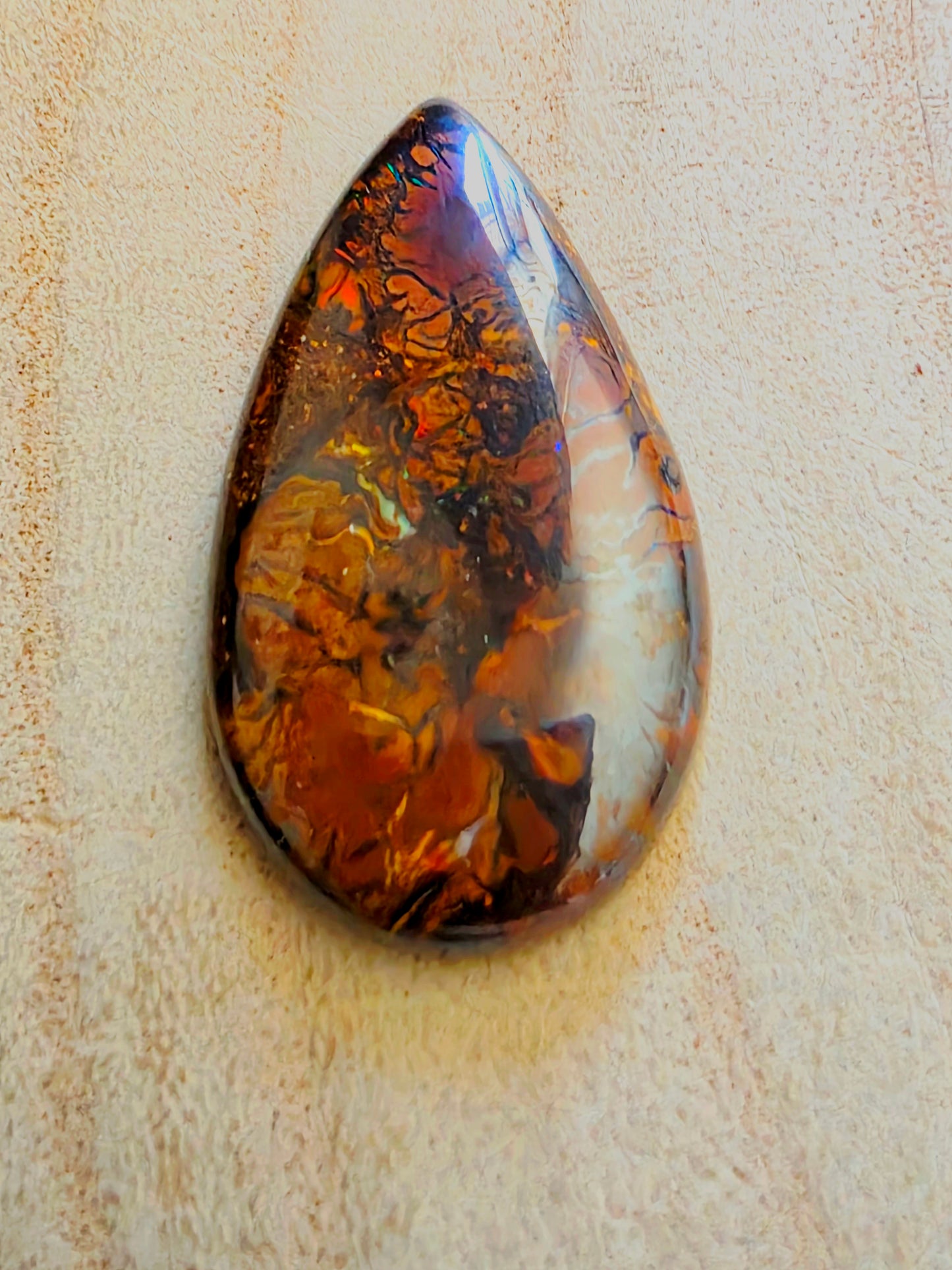 A teardrop-shaped polished opal cabochon with a marbled pattern of amber orange, deep brown, black, and white, displayed on a textured beige background.