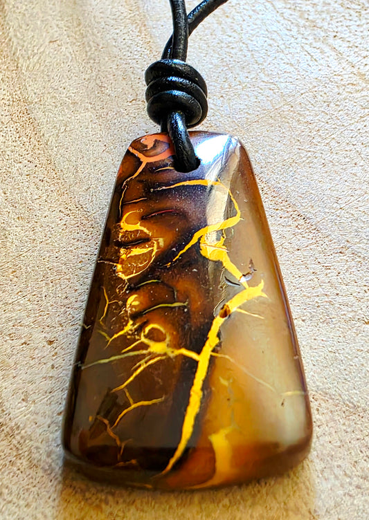 A polished brown stone opal with yellow veining, suspended from a black cord, shown on a textured beige surface.