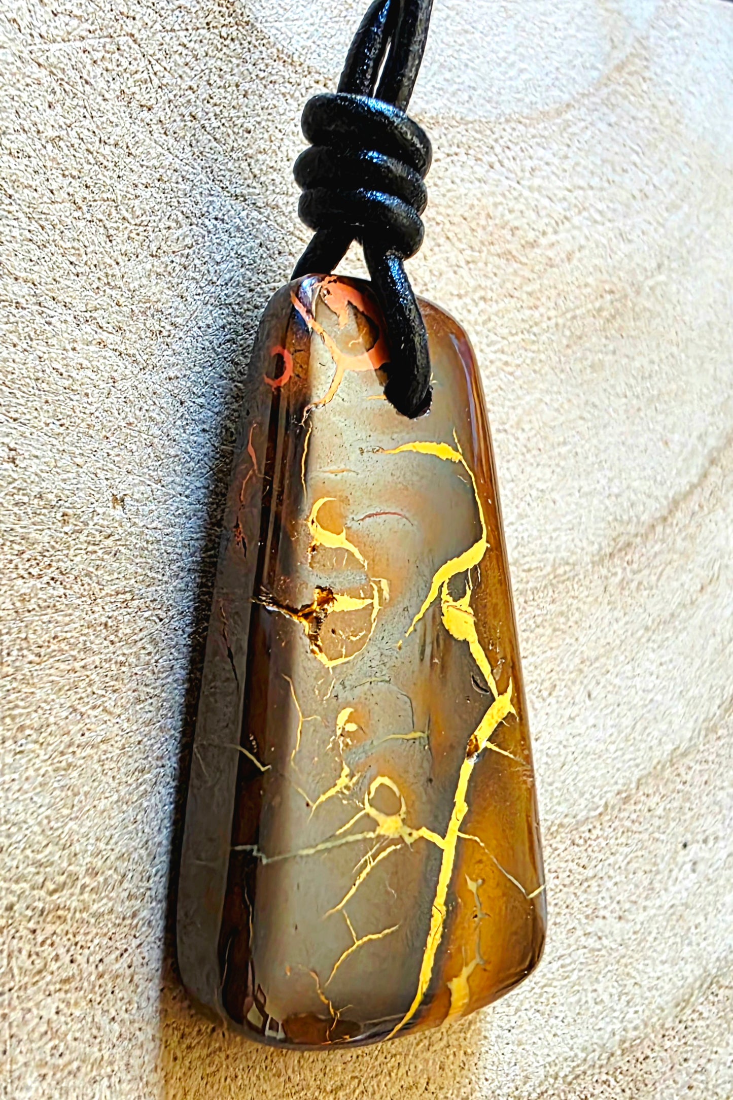 A polished teardrop-shaped stone pendant with a brown, amber, and gold marbled pattern, suspended from a black cord on a textured beige background.