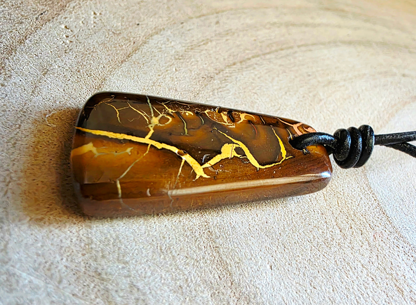 A polished brown stone pendant with striking gold veining, threaded onto a black cord, resting on a light beige textured surface.