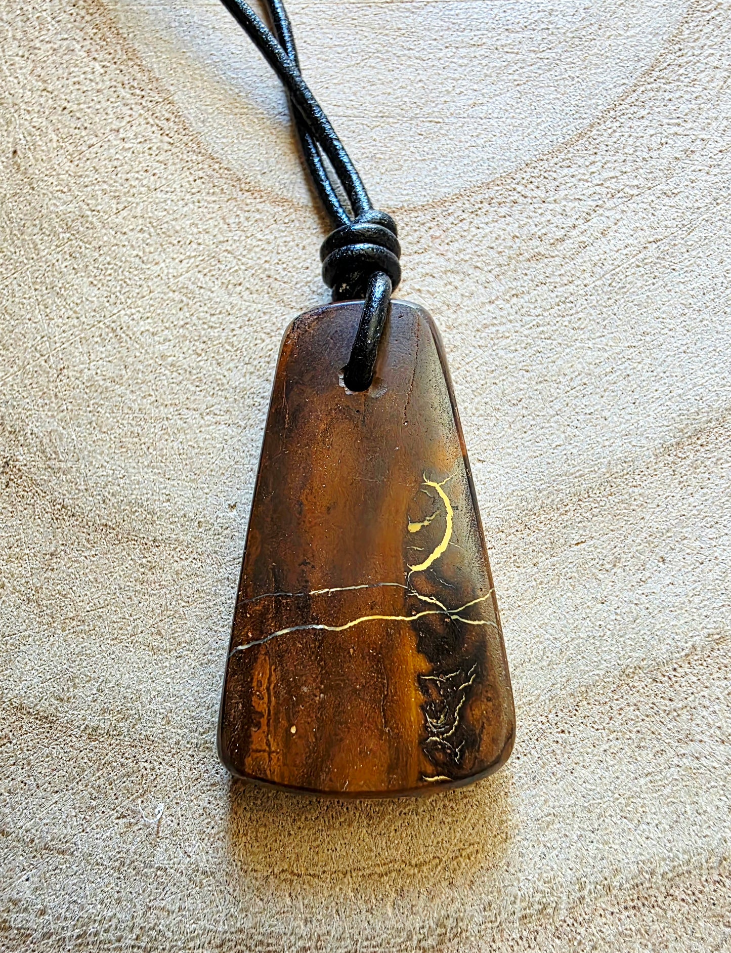 A brown polished stone pendant with natural inclusions and a black cord necklace, displayed on a textured beige surface.