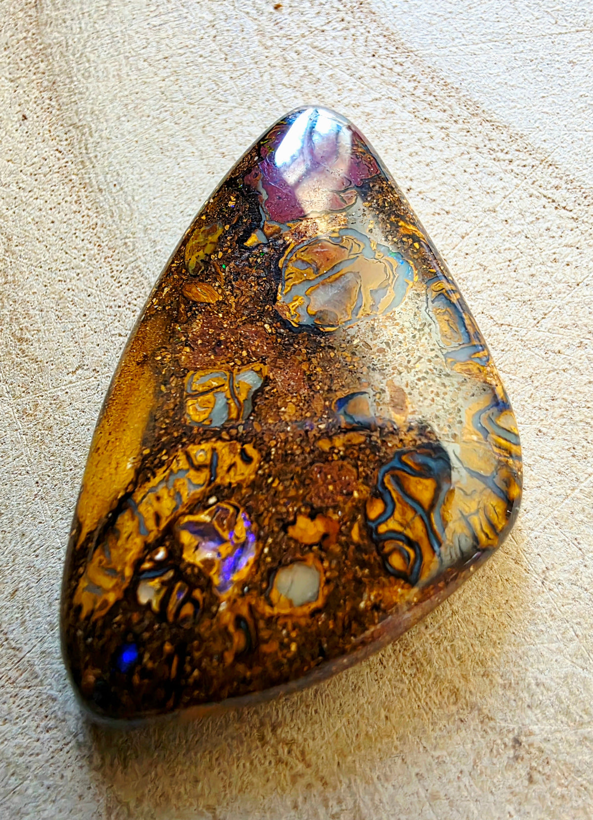 A polished teardrop-shaped opal stone with swirling bands of gold, brown, black, and iridescent blue-purple flashes, resting on a textured beige surface.
