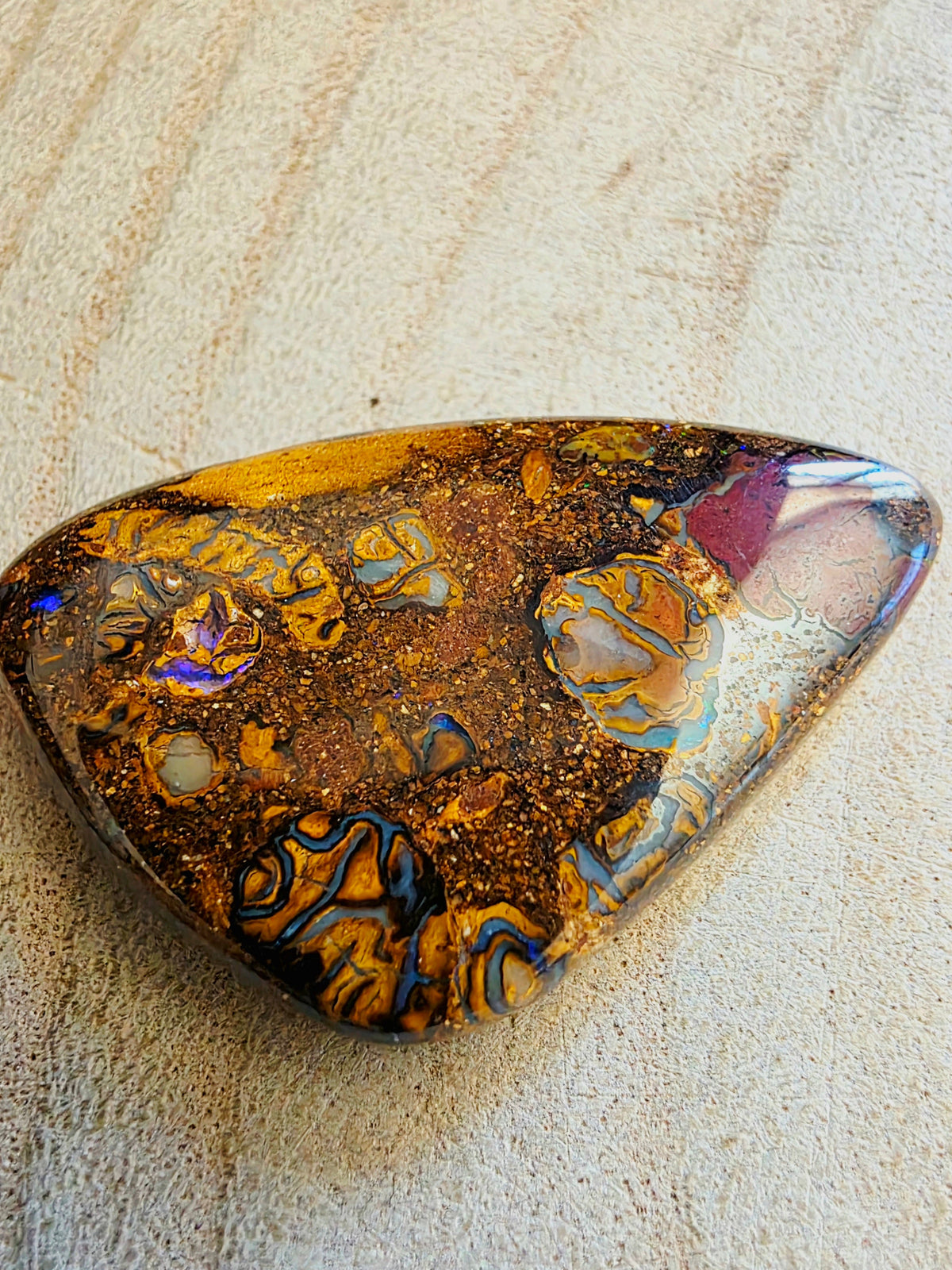 A polished teardrop-shaped opal stone with intricate patterns of gold, brown, orange, blue, and purple, resting on a textured beige surface.