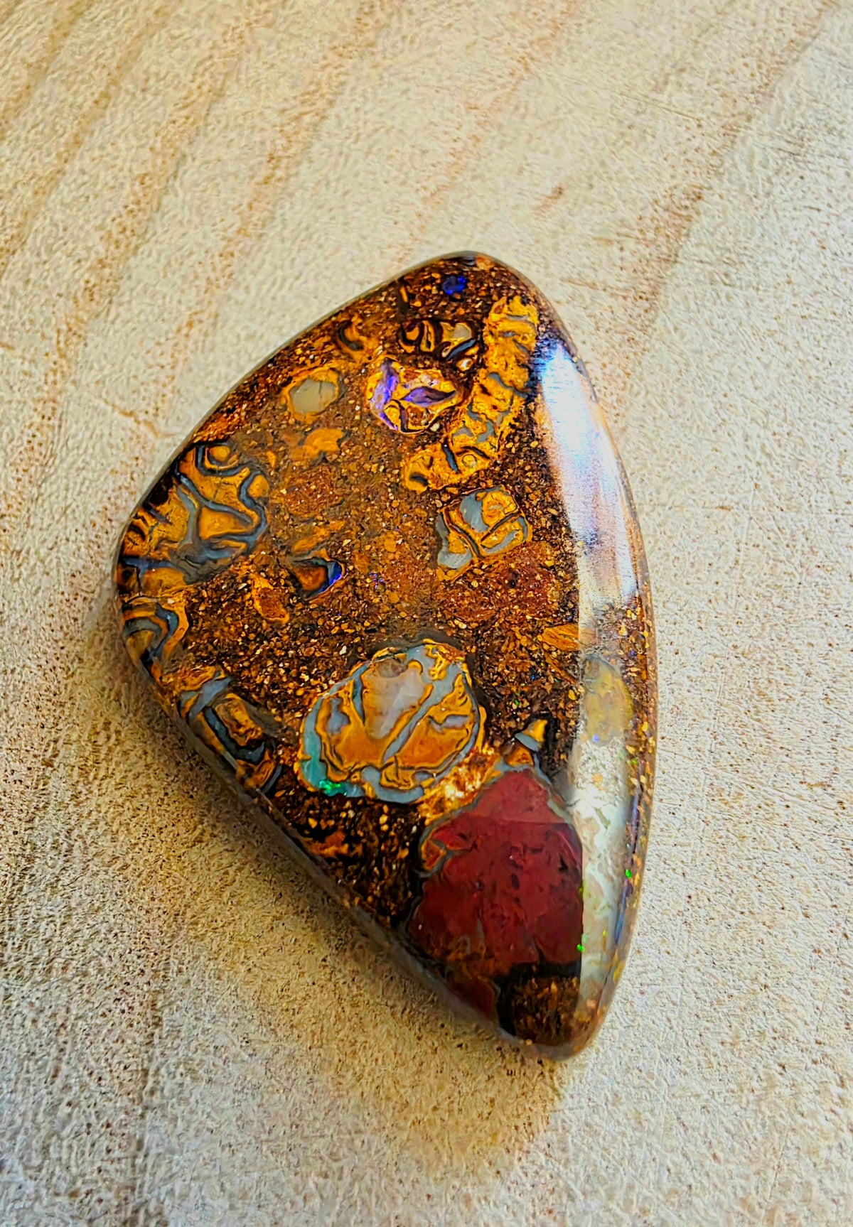 A polished boulder gemstone with a rich pattern of gold, orange, red, and iridescent blue flashes, resting on a textured beige wood surface.