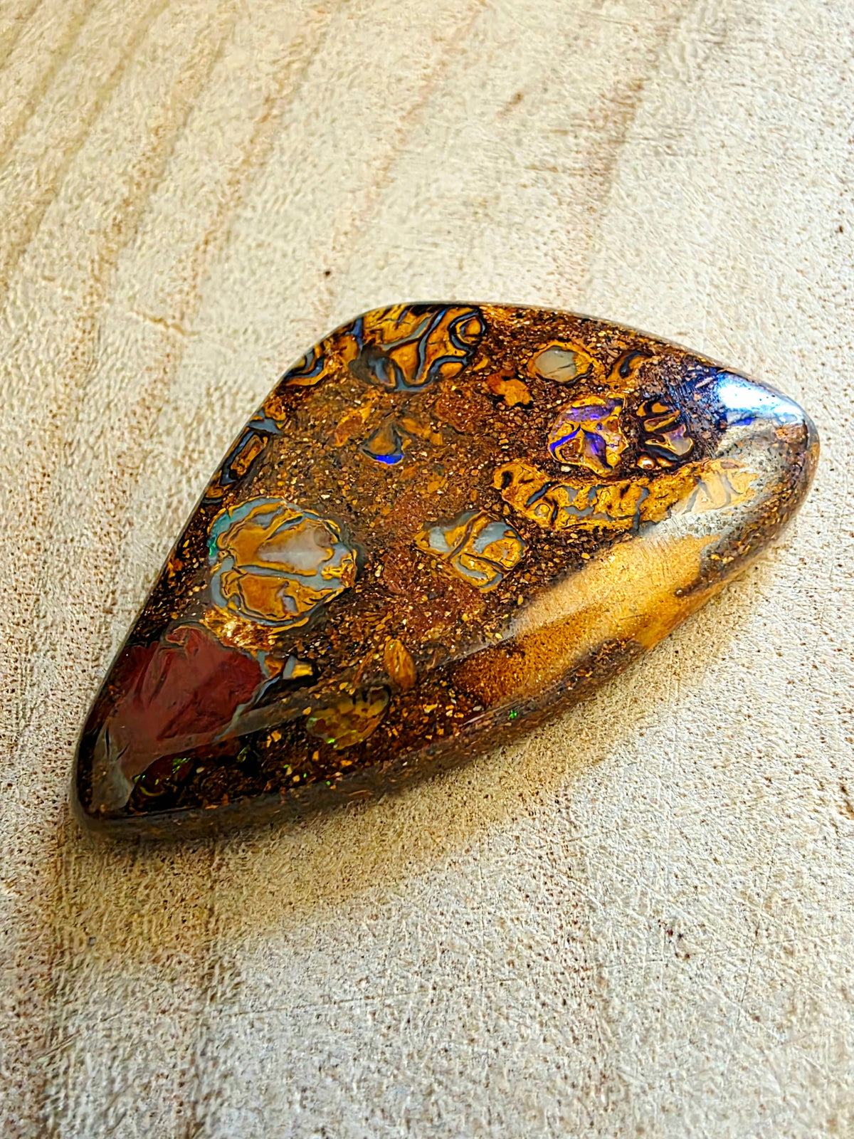 A polished natural bohemian gemstone with intricate patterns of gold, orange, brown, and iridescent blue-purple hues, resting on a textured beige surface.