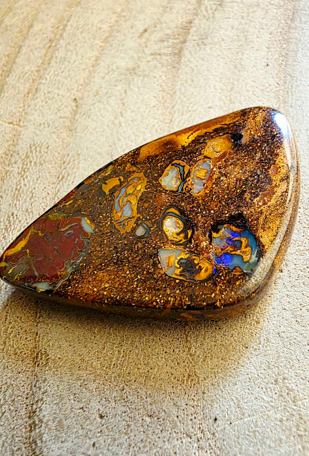 A polished natural opal stone with a teardrop shape, featuring a mottled pattern of golden-brown, orange, red, and iridescent blue flashes, resting on a textured beige surface.
