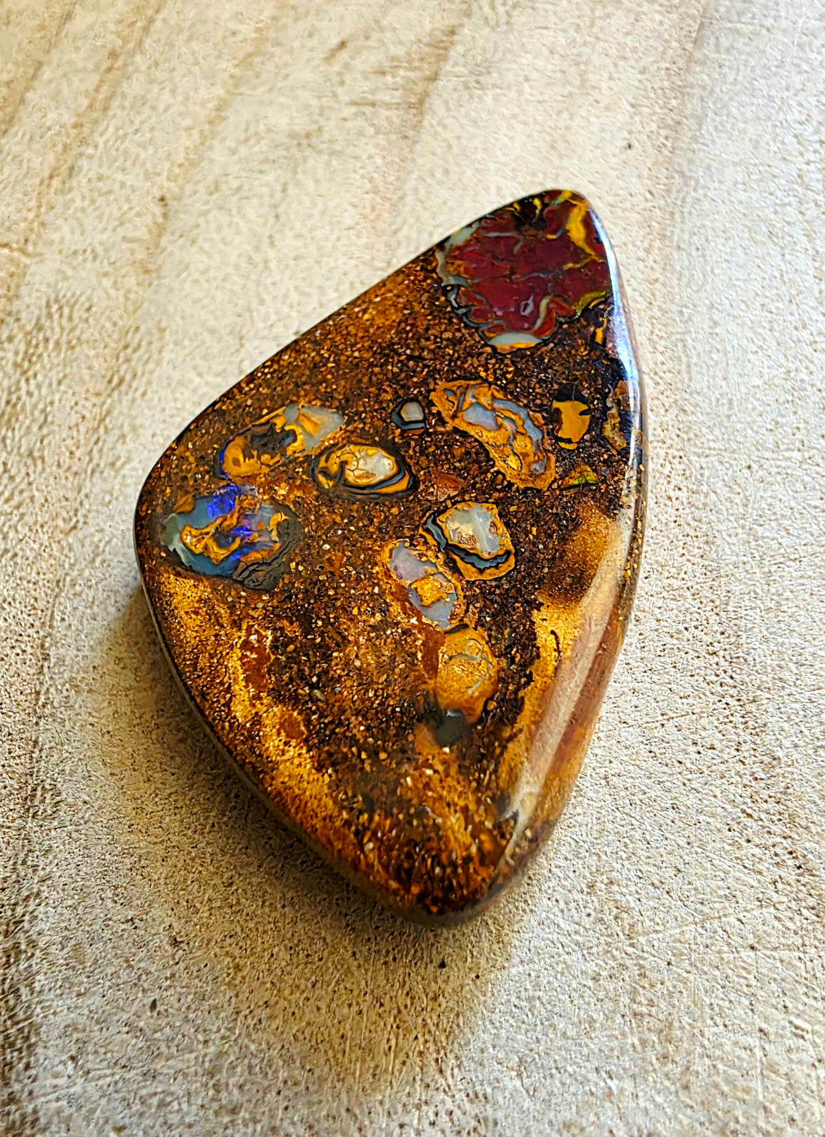 A polished opal stone with intricate patterns of gold, brown, orange, blue, and purple, resting on a textured beige surface.