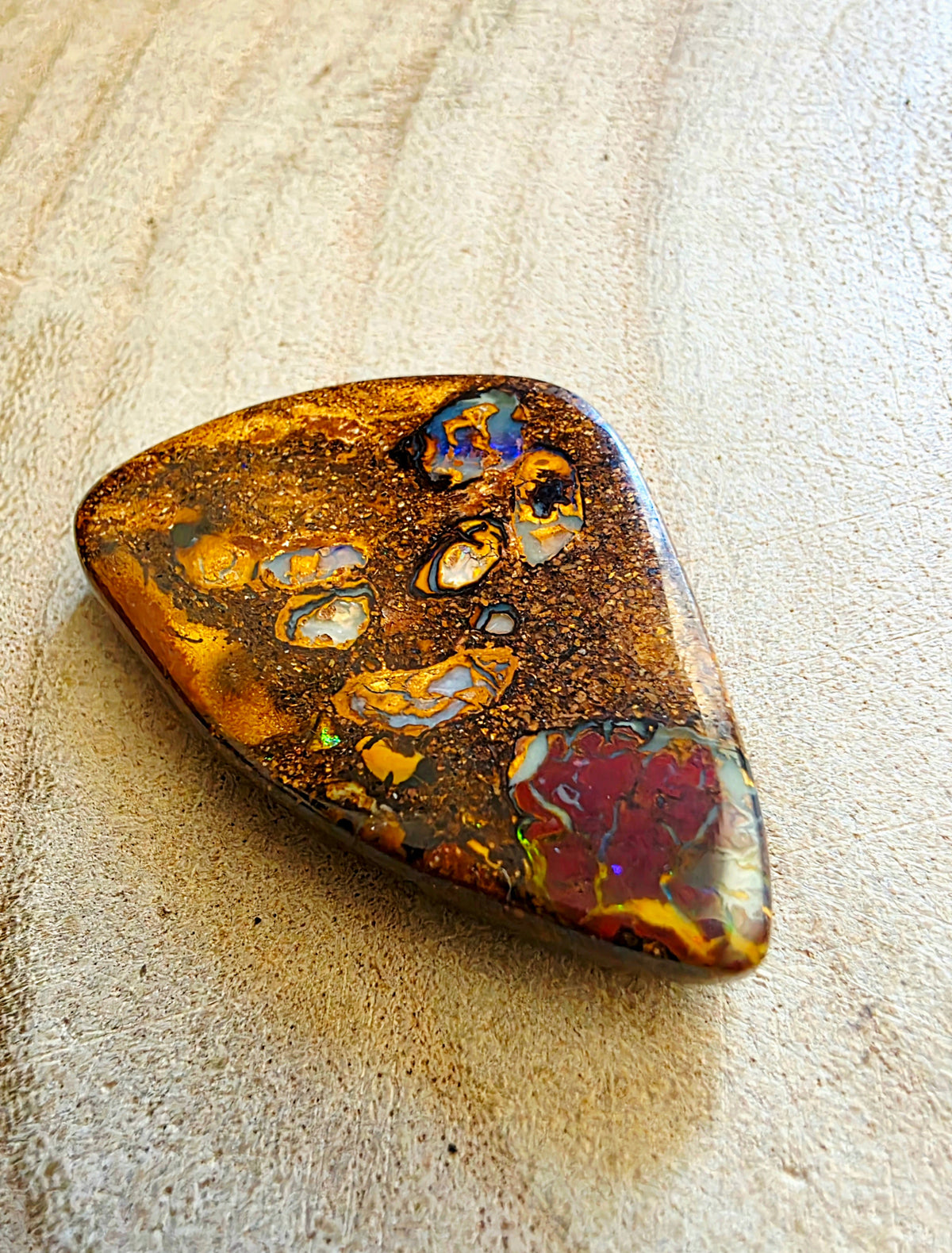 A polished teardrop-shaped opal stone with a rich pattern of gold, brown, red, and iridescent blue flashes, resting on a textured beige surface.