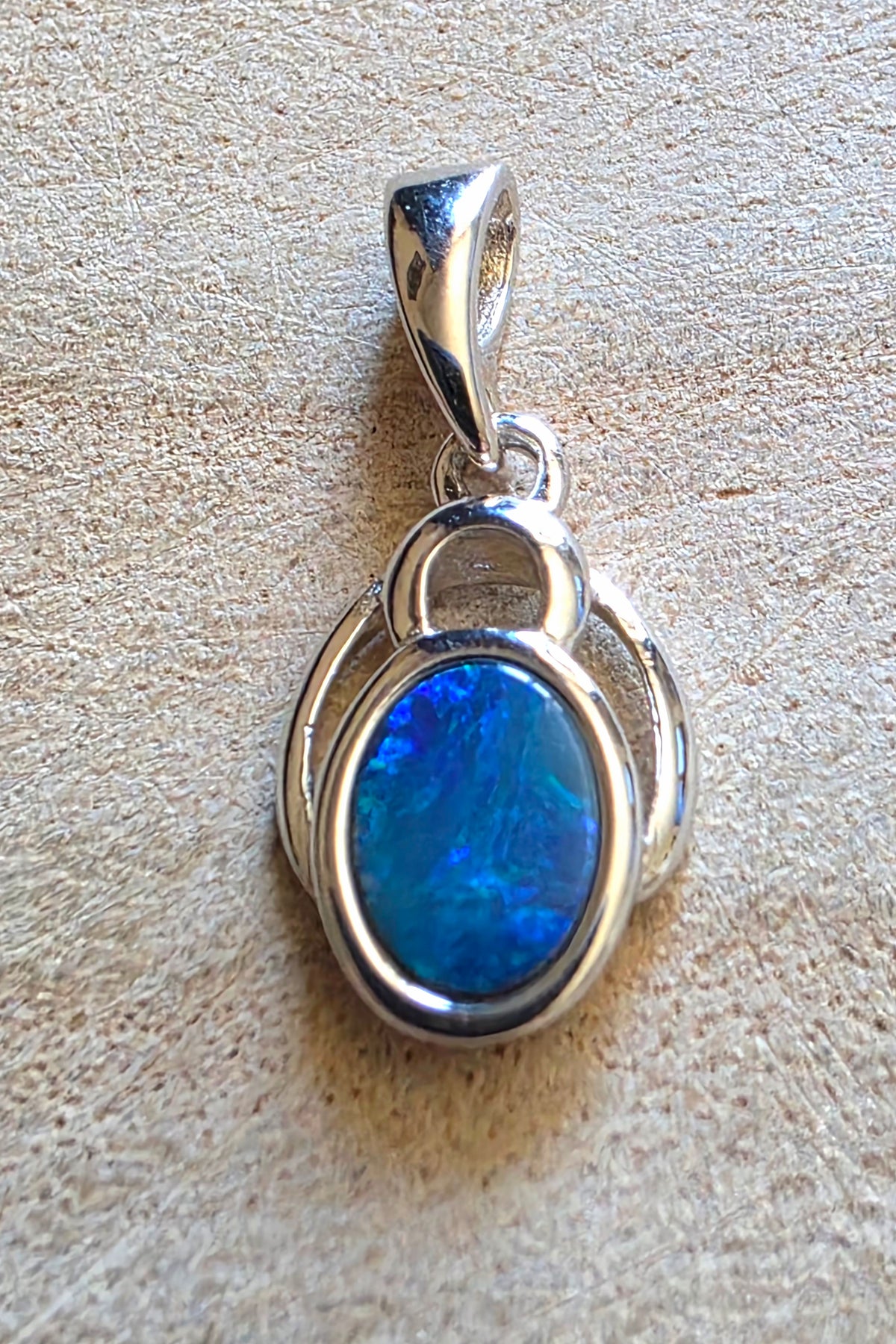 A sterling silver pendant featuring an oval-cut blue opal gemstone, displayed on a textured beige surface.
