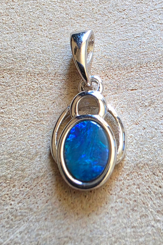 A sterling silver pendant featuring an oval-cut blue opal gemstone, displayed on a textured beige surface.