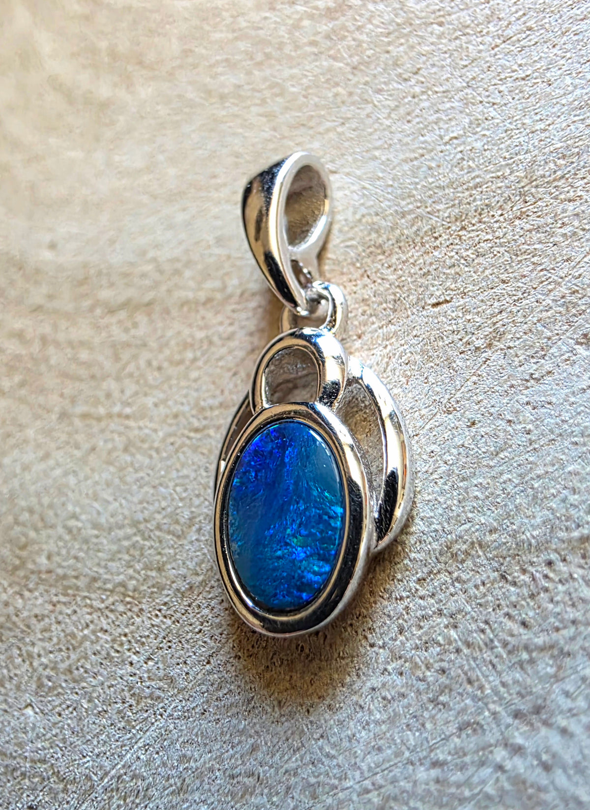 A silver pendant featuring an oval blue opal stone, displayed on a textured beige background.
