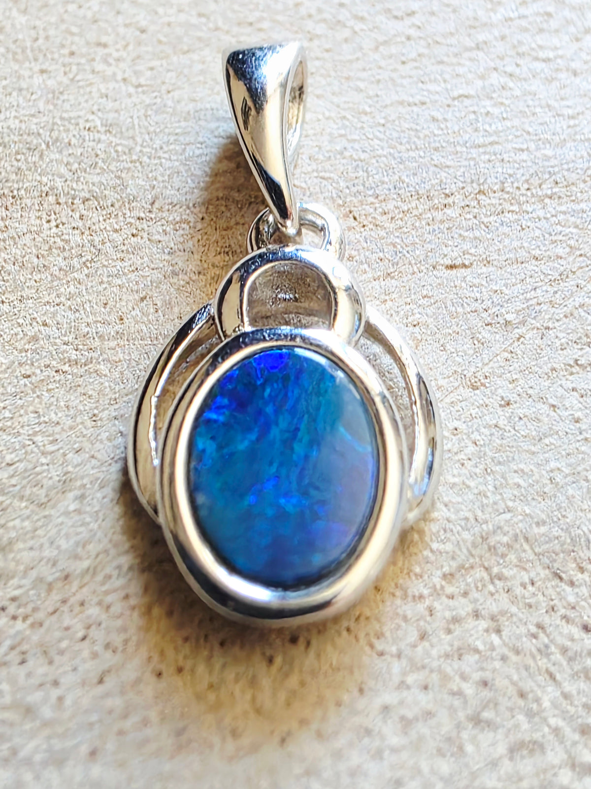 A sterling silver pendant featuring an oval blue opal gemstone, displayed on a textured beige surface.