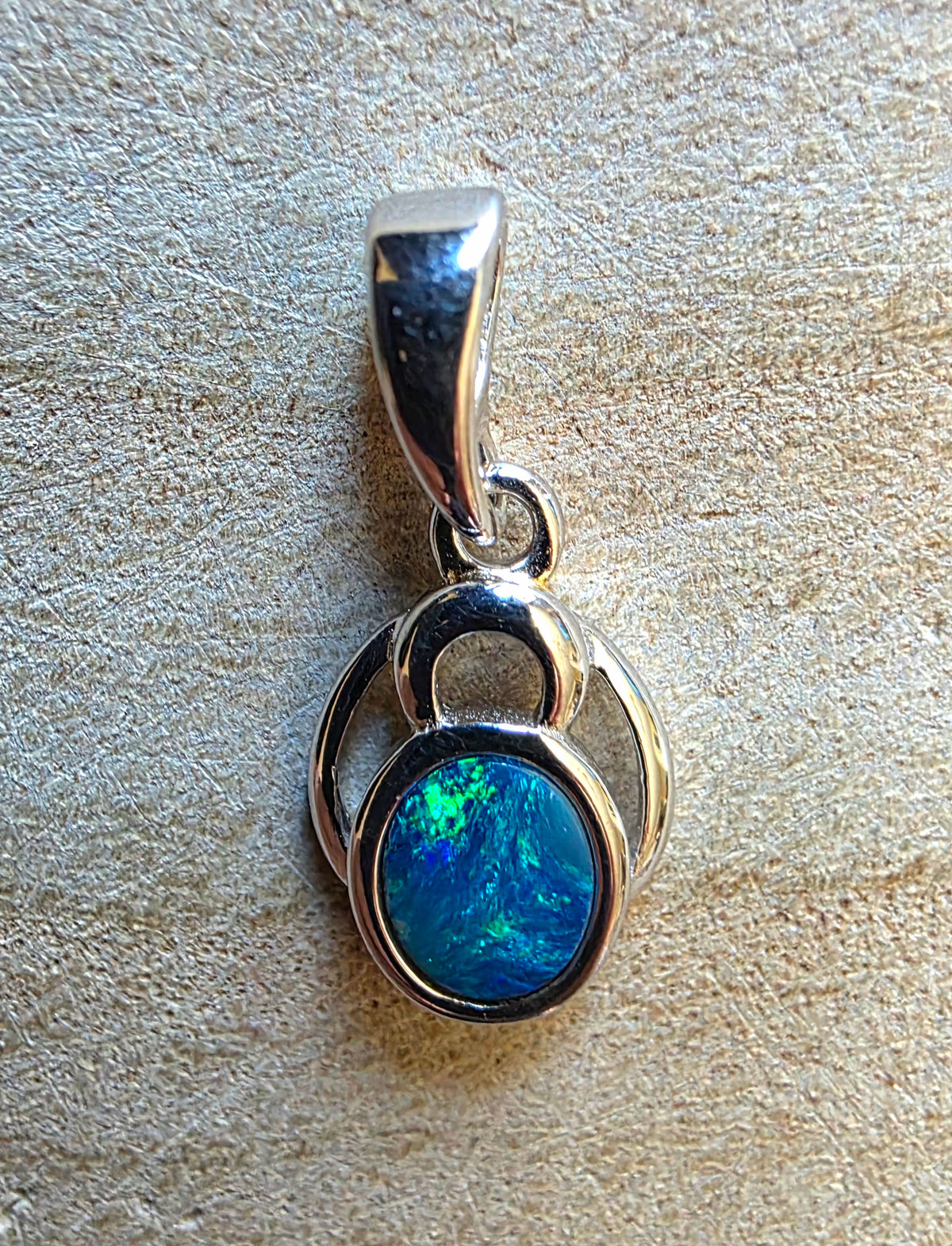 A sterling silver pendant featuring a round, vibrant blue and green opal gemstone, displayed on a textured beige surface.
