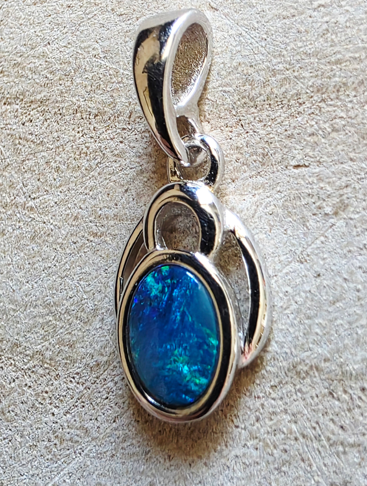 A sterling silver pendant featuring an oval blue opal gemstone, displayed on a textured beige background.