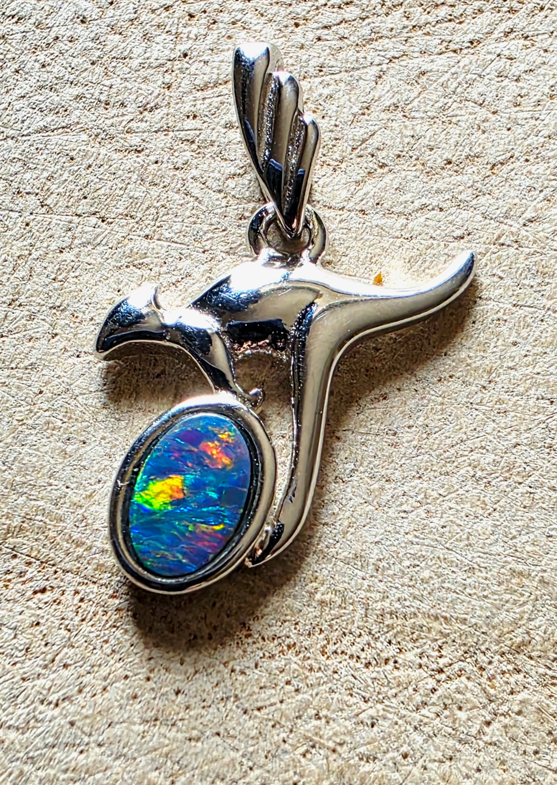 A sterling silver pendant shaped like a kangaroo, featuring a round iridescent opal stone with blue, green, yellow, and orange play-of-colour, set against a textured beige background.