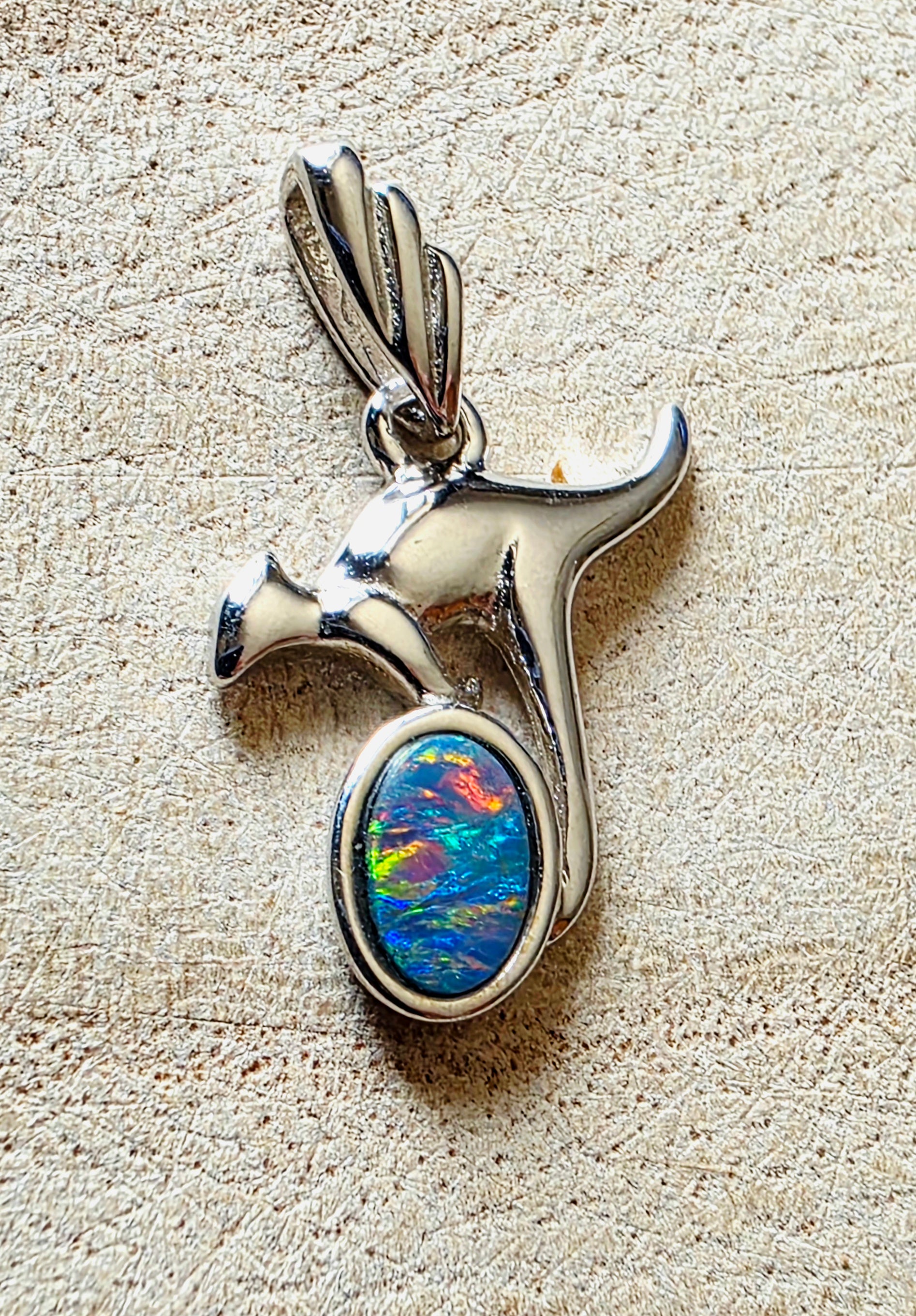 A sterling silver pendant shaped like a kangaroo with a colourful, iridescent opal gemstone in an oval bezel setting, displayed on a textured beige surface.