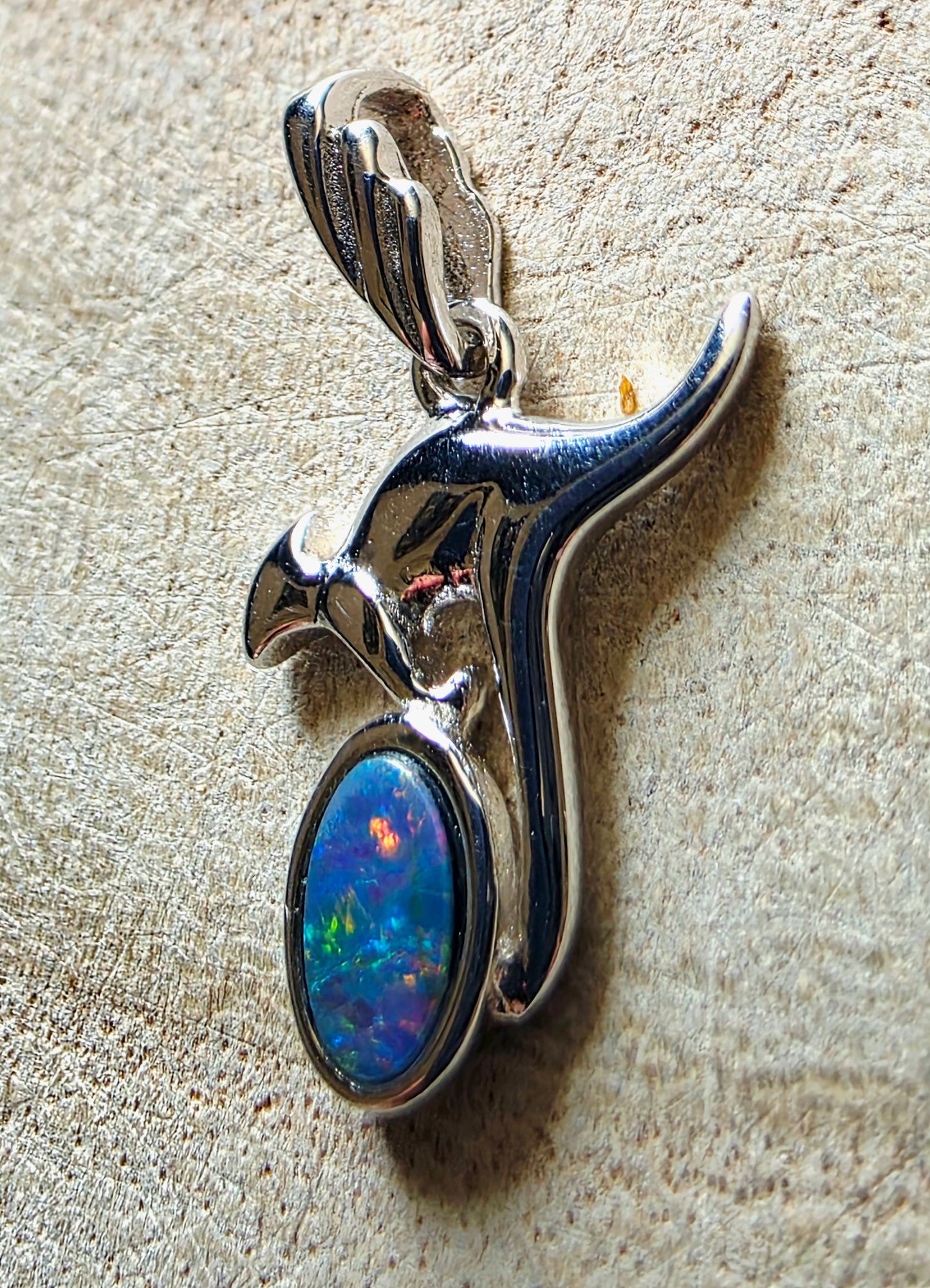 A sterling silver pendant shaped like a kangaroo, featuring a round iridescent opal stone with blue, green, and orange flashes, displayed on a textured beige surface.