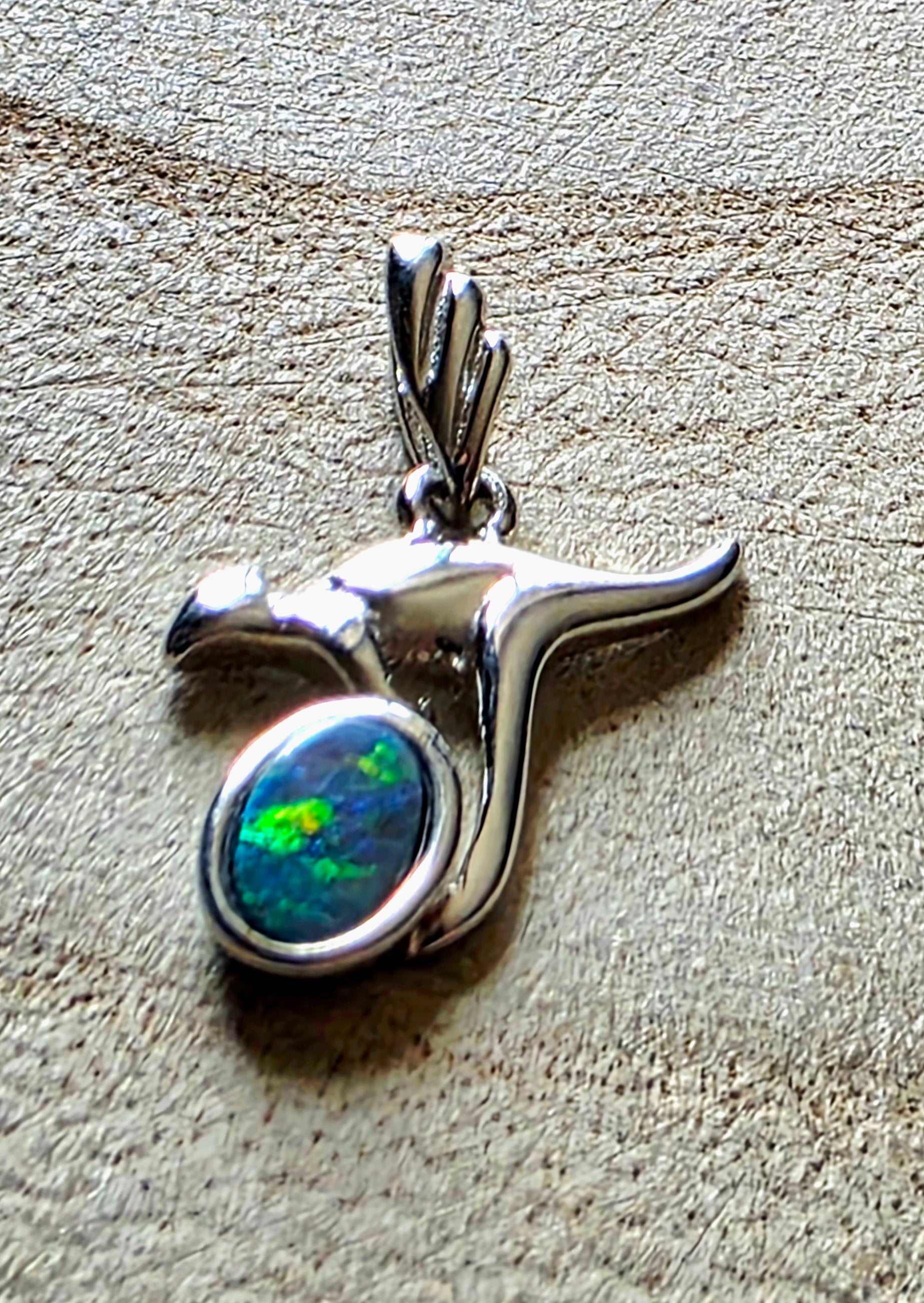 A sterling silver pendant shaped like a kangaroo, featuring a round bezel-set iridescent opal with green and blue flashes, shown on a textured beige surface.