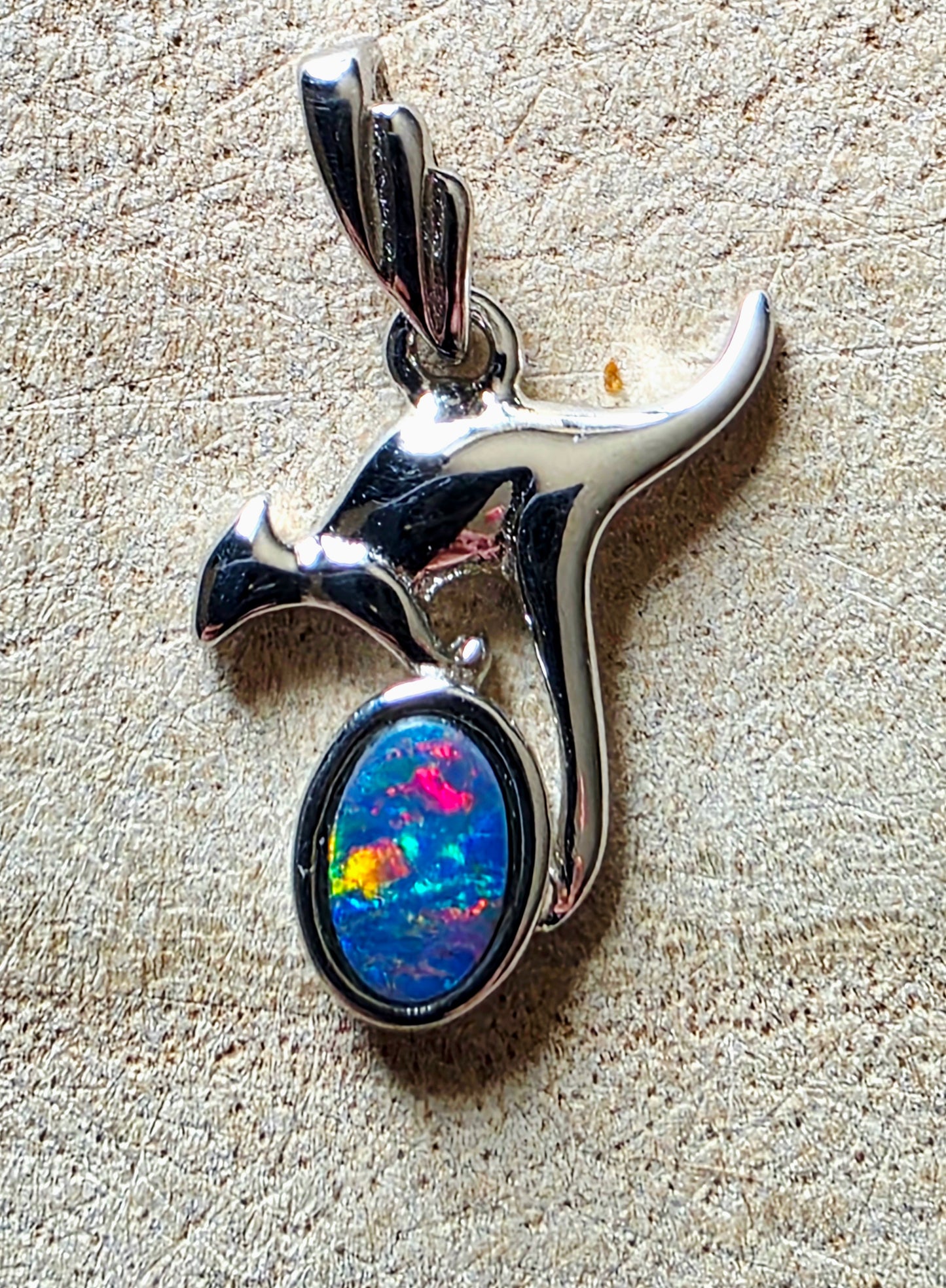 A polished silver pendant shaped like a kangaroo, featuring an oval iridescent opal stone with vivid blue, pink, yellow, and green flashes, set against a textured beige background.