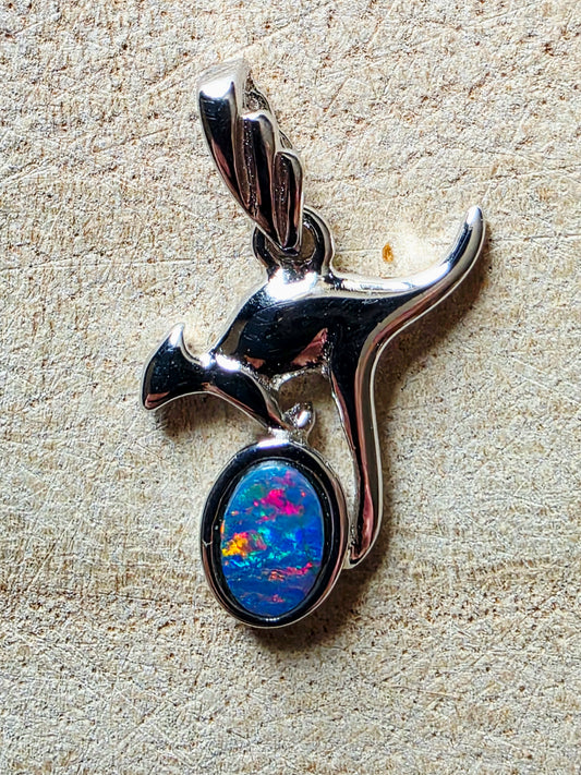A polished silver pendant shaped like a kangaroo, featuring an oval inset stone with vivid blue, purple, orange, and pink opal-like play-of-colour, photographed on a textured beige surface.