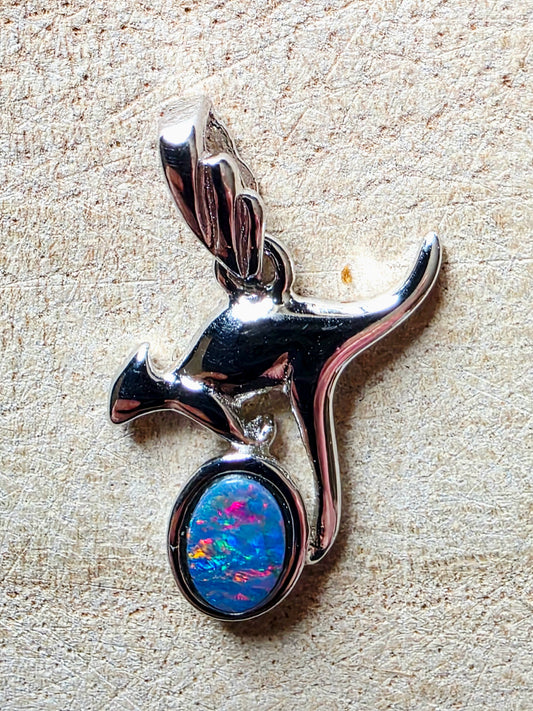 A polished silver pendant shaped like a kangaroo, featuring an oval cabochon stone with vivid blue, pink, and orange iridescent opal-like colours, photographed on a textured beige surface.