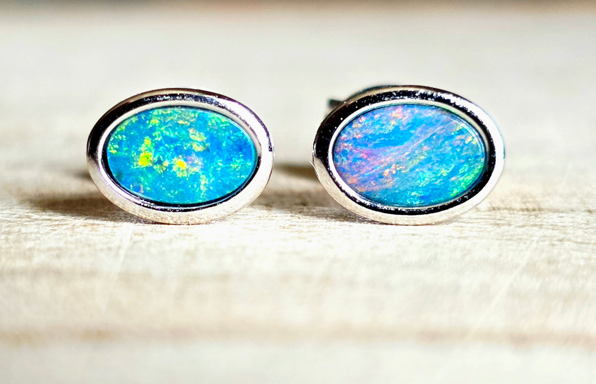 A pair of oval stud earrings with vibrant blue and green opal inlays set in silver, resting on a light wood surface.