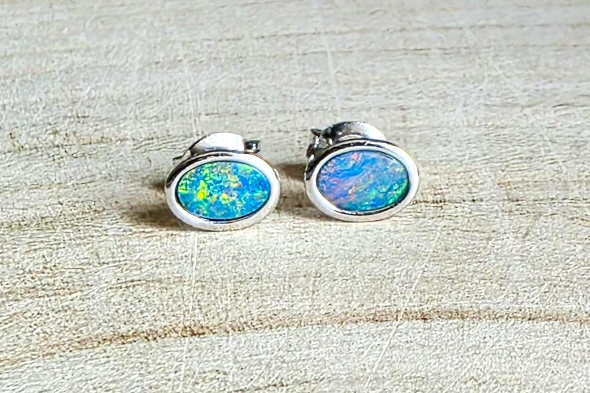 A pair of oval stud earrings with vibrant blue, green, and orange iridescent opal inlays set in silver, resting on a light beige textured wood surface.