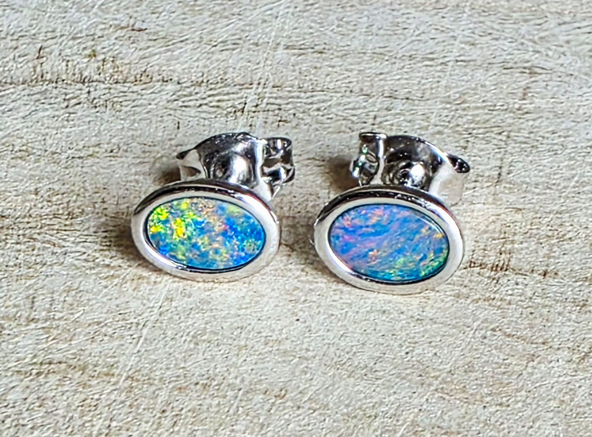 A pair of oval stud earrings with iridescent blue, green, and yellow opal inlays set in polished silver, displayed on a textured beige surface.