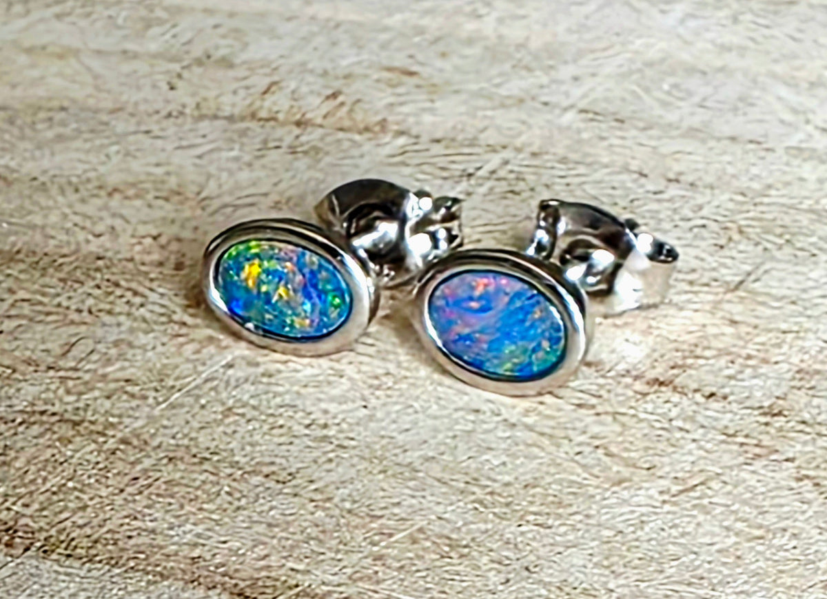 A pair of oval stud earrings with iridescent blue, green, and orange opal inlays set in silver, resting on a light wood surface.