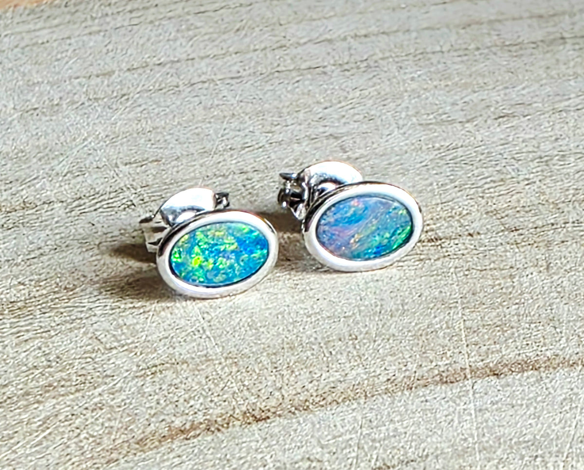 A pair of oval stud earrings with vibrant blue and green opal inlays set in silver, resting on a light beige textured surface.