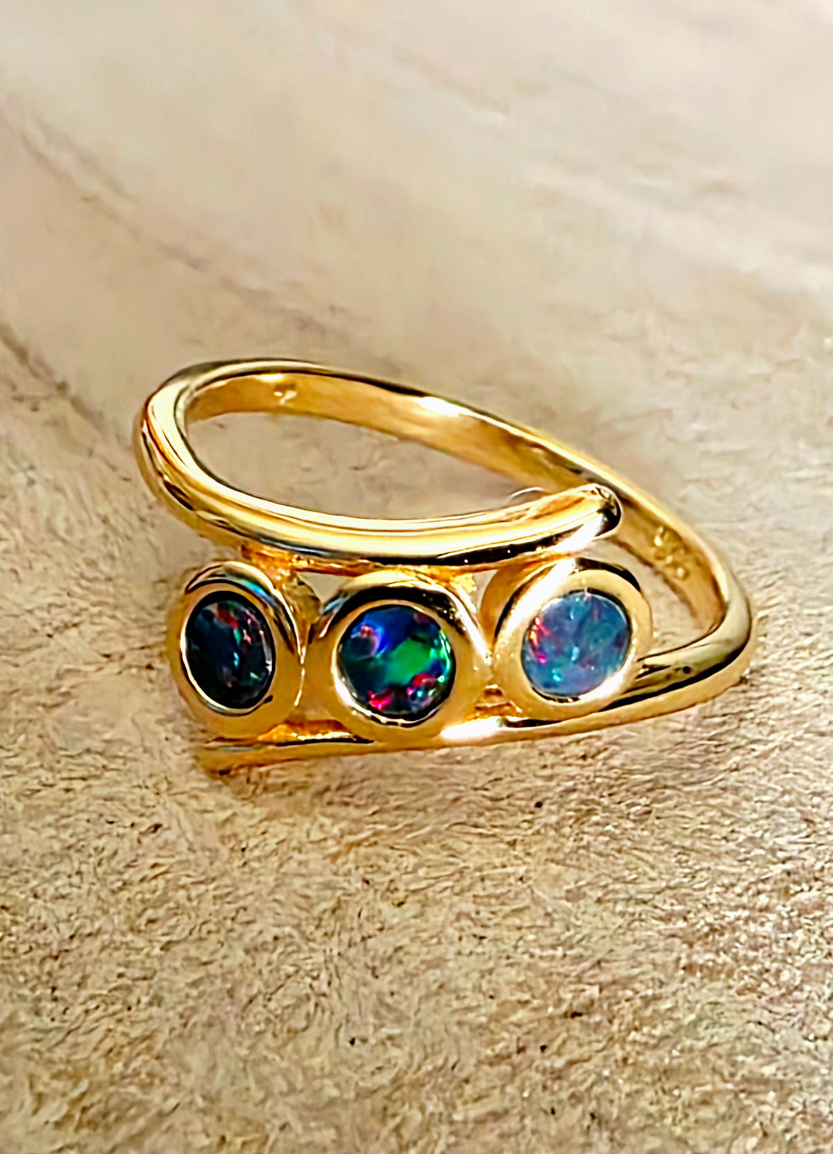 A gold ring featuring three round bezel-set opal stones, displayed on a textured beige background.