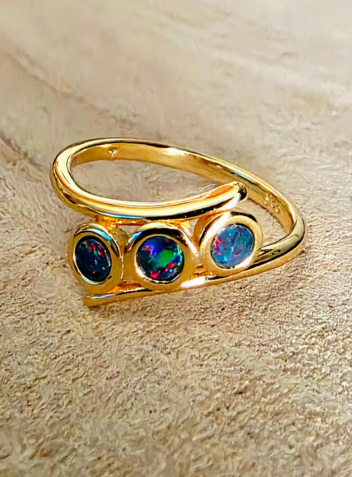 A gold ring with three round iridescent opal gemstones, displayed on a textured beige background.
