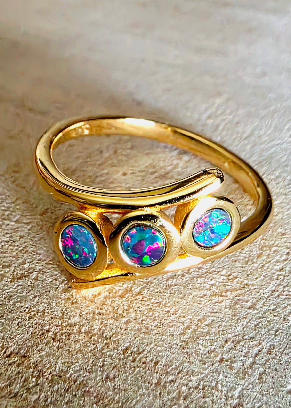 A gold ring featuring three round bezel-set iridescent opal stones, displayed on a textured beige background.