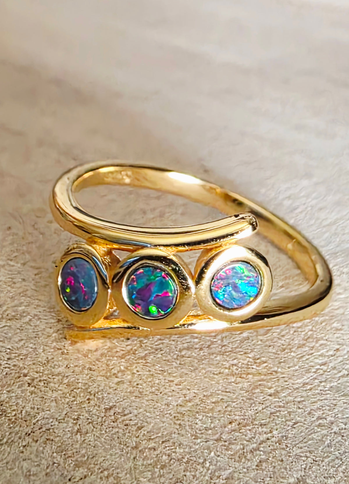 A gold ring featuring three round bezel-set iridescent opal stones, displayed on a textured background.