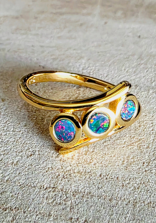 A gold ring featuring three round bezel-set iridescent opal stones, displayed on a textured beige fabric background.