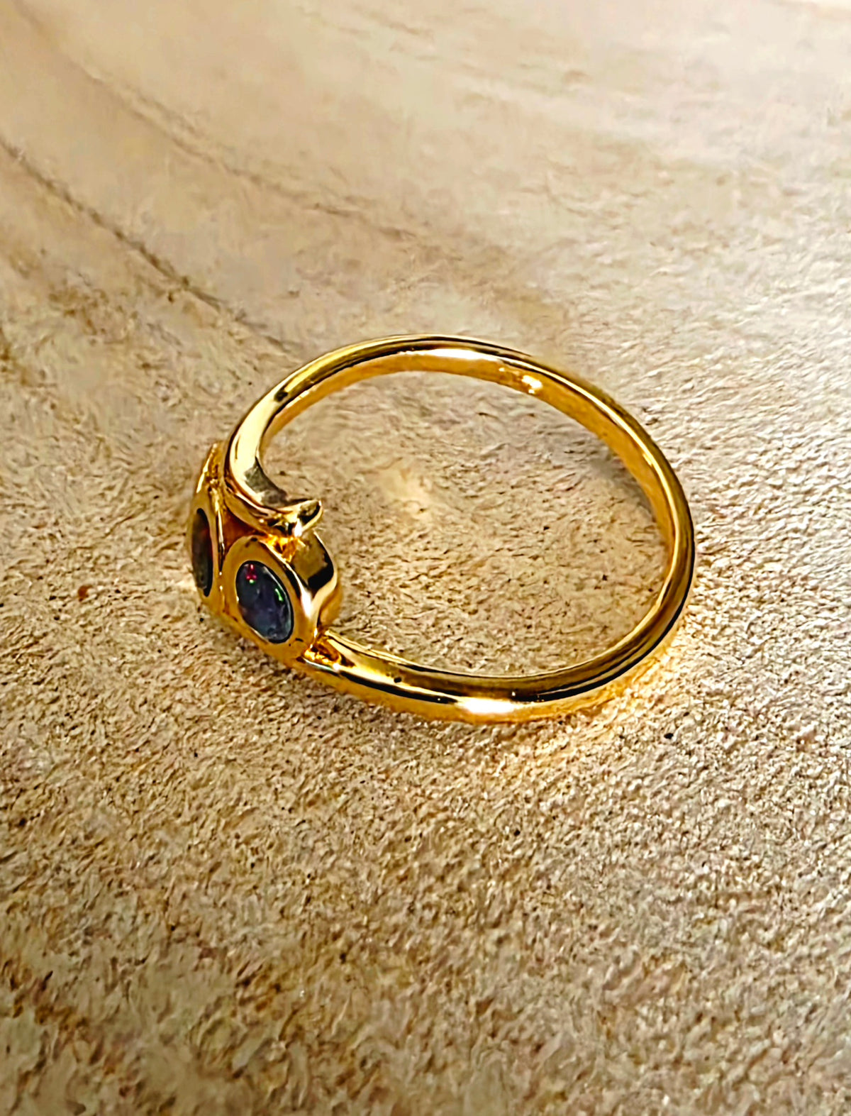 A gold ring with three round bezel-set iridescent opal stones, displayed on a textured beige background.