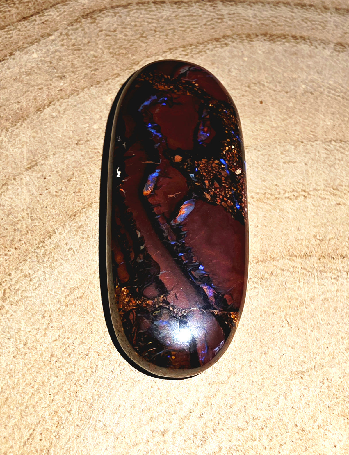 A polished oval cabochon opal with a deep reddish-brown body and vivid iridescent blue, purple, and gold inclusions, photographed on a textured beige surface.