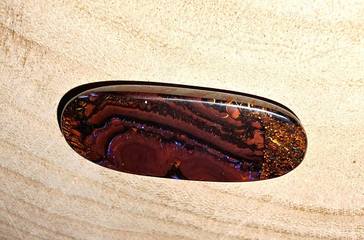 An oval opal with a polished boulder opal pattern in deep red, brown, and black, embedded in a textured beige plaster ceiling.