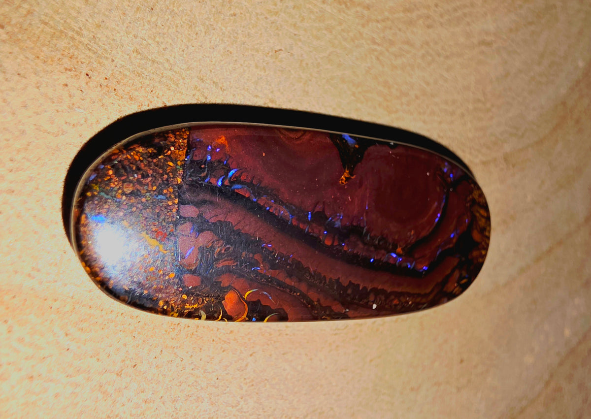 A polished oval cabochon opal with a marbled pattern of deep red, black, and iridescent blue-orange flashes, set in a black bezel against a textured beige background.