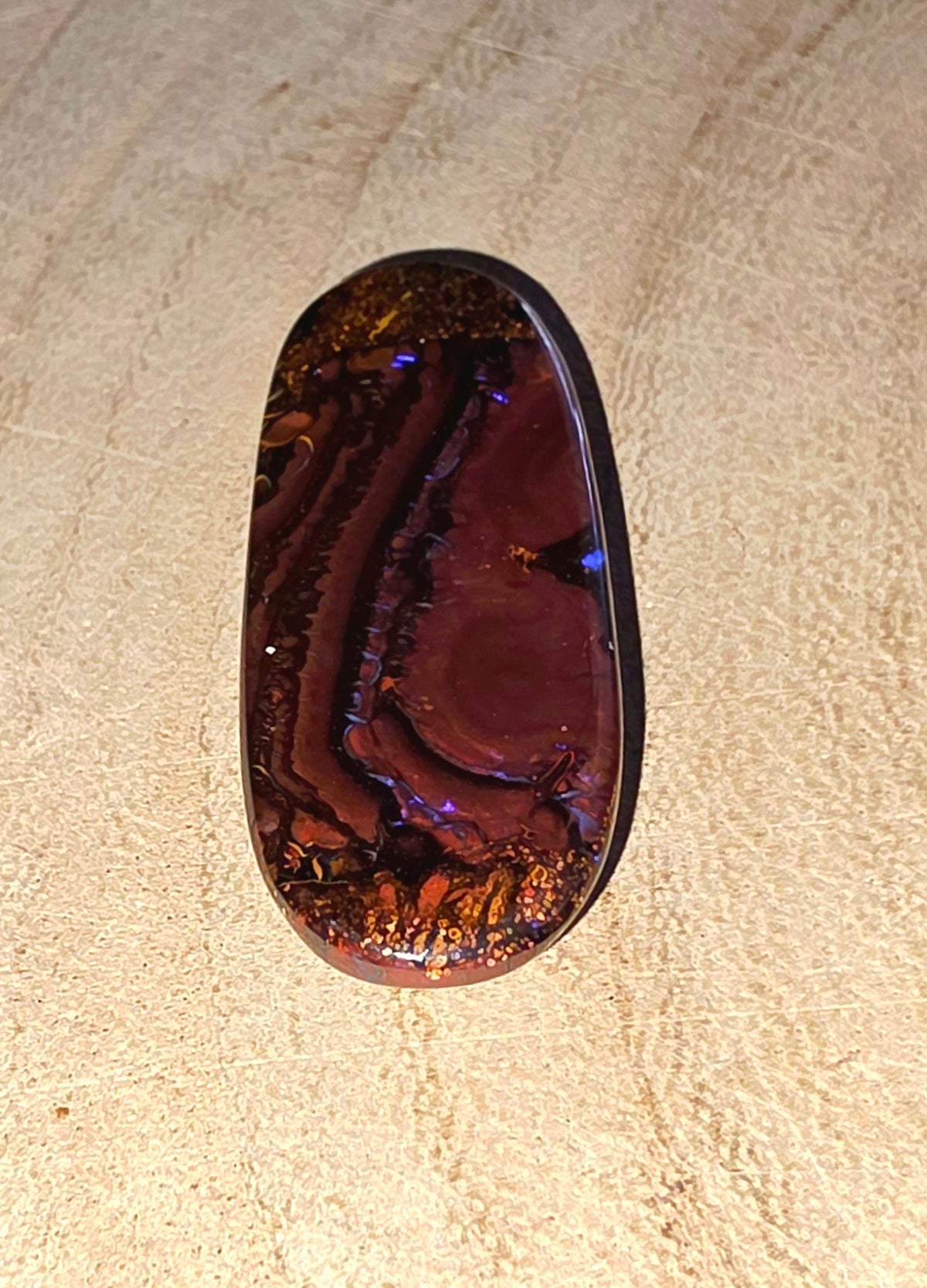 A polished oval cabochon opal with a deep reddish-brown body and iridescent play-of-colour, including blue and orange flashes, resting on a textured beige surface.