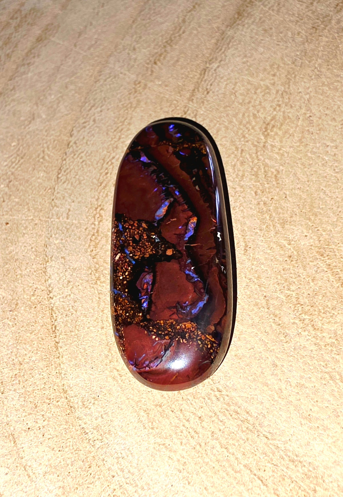 A polished oval cabochon opal with a deep reddish-brown body and vivid iridescent blue, purple, and orange flashes, displayed on a textured beige surface.