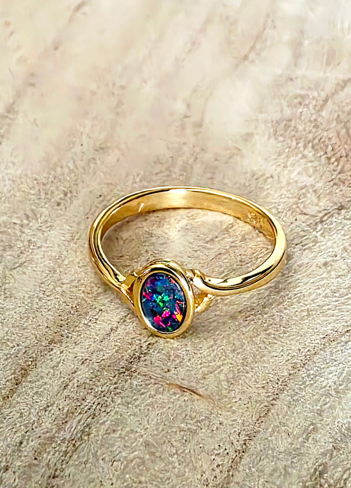 A gold ring featuring an oval multicolor opal gemstone, resting on a textured beige surface.