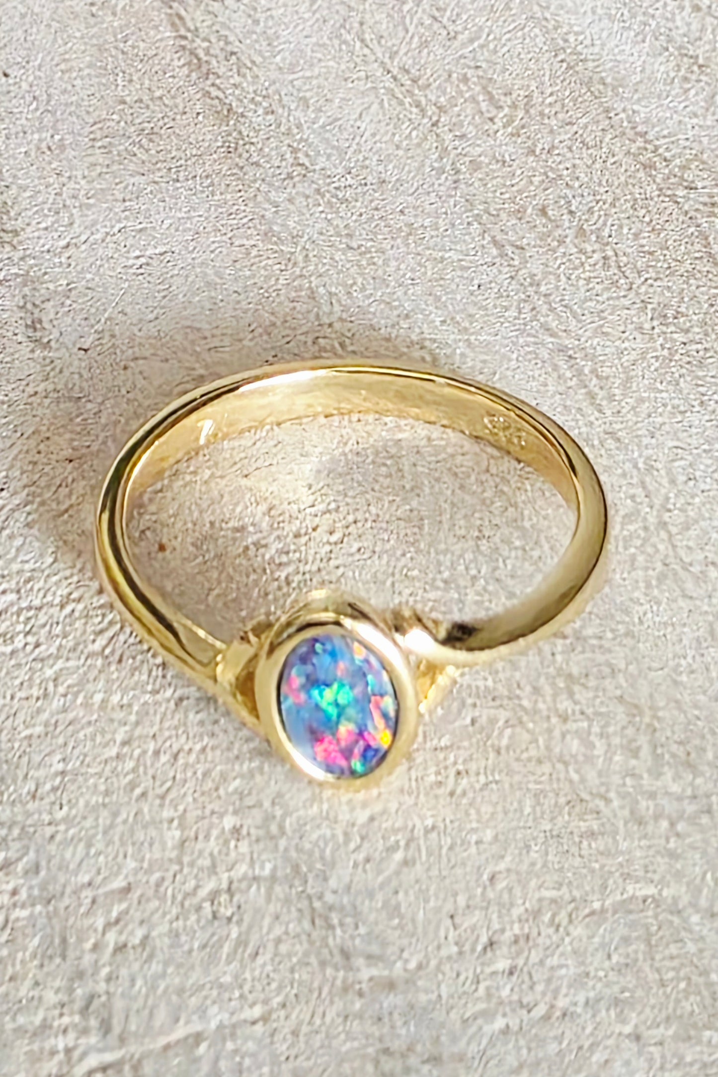 A gold ring featuring an oval-cut iridescent opal gemstone with vibrant blue, green, and pink flashes, set against a textured beige background.