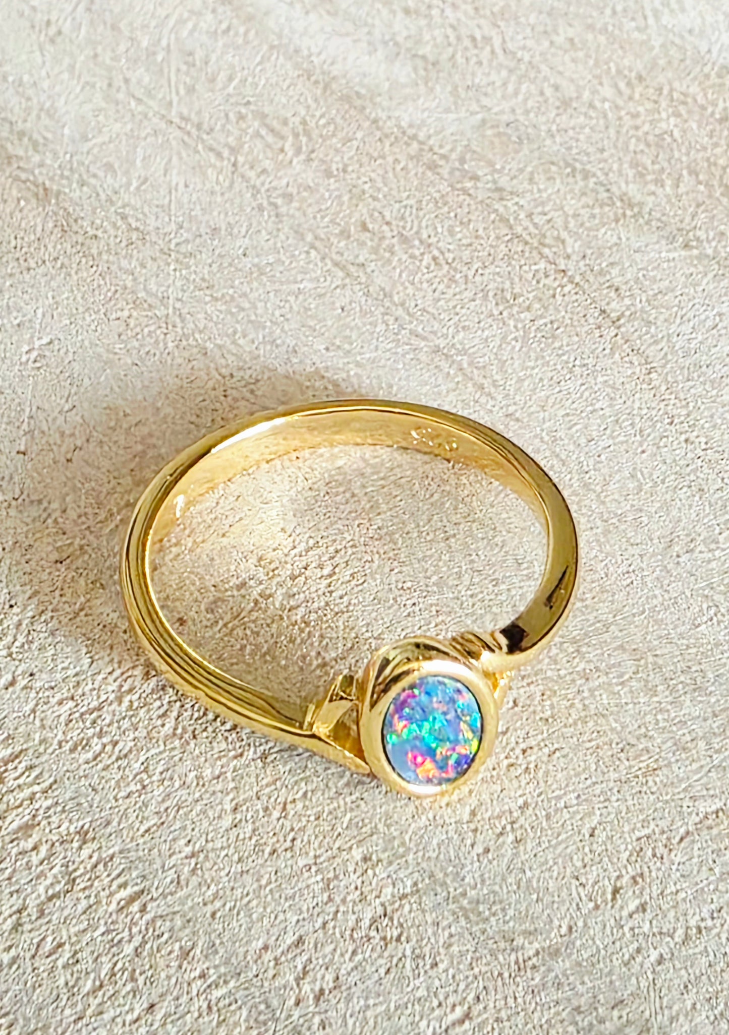 A gold ring featuring an oval-shaped iridescent opal gemstone, displayed on a textured beige fabric background.