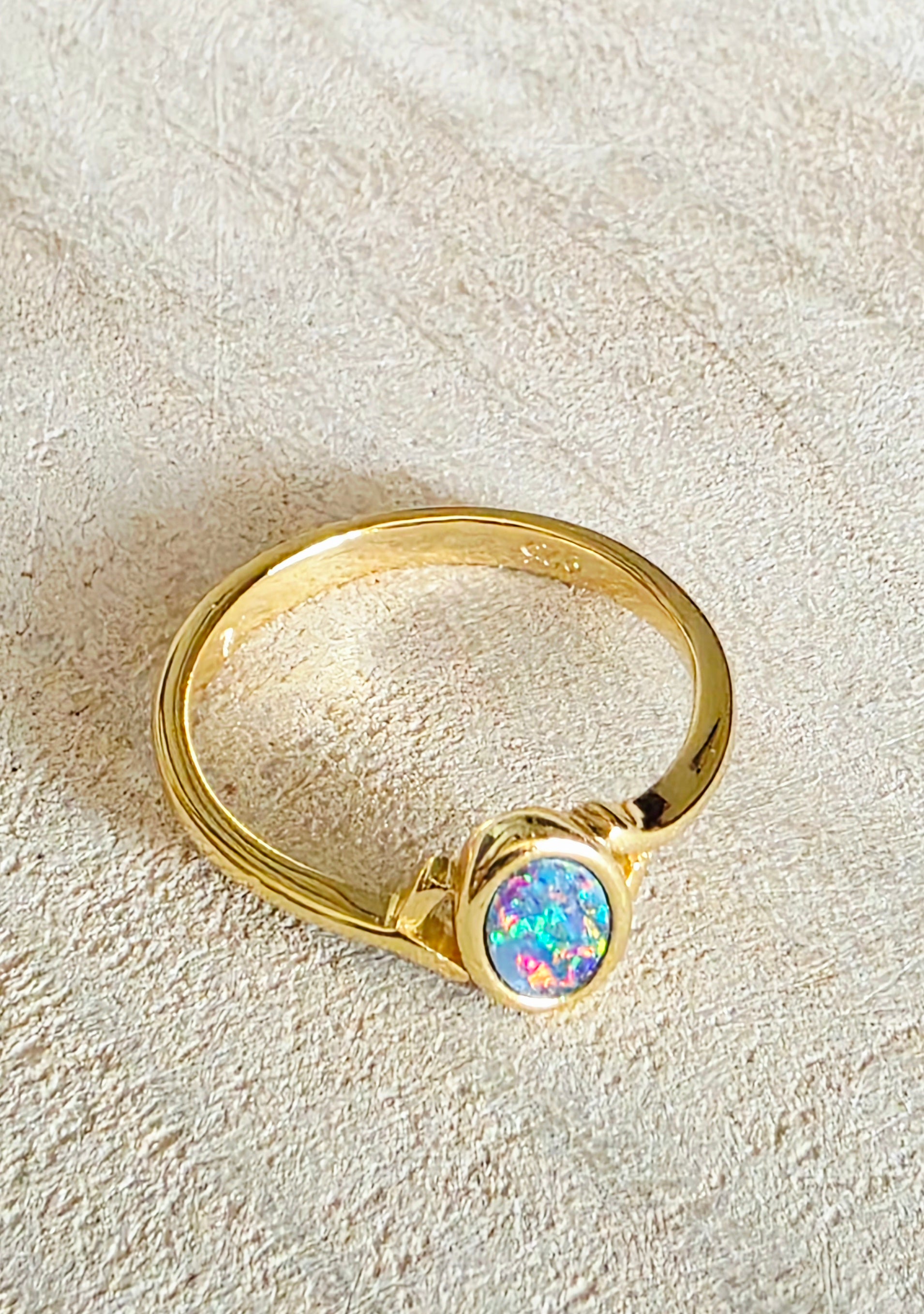 A gold ring featuring an oval-shaped iridescent opal gemstone, displayed on a textured beige fabric background.